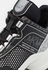 Baskets noires et grises avec empiècements en maille, accents en cuir texturé, matériel réfléchissant et étiquette de marque sur le côté.