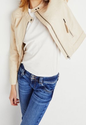 Giacca cropped in pelle beige con zip e bottoni a pressione, abbinata a una t-shirt bianca e jeans fitted blu scuro. Composizione di abbigliamento casual.