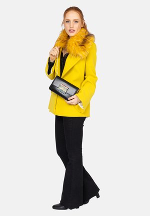 Gul frakke med faux fur krave, parret med en sort top og flagrende sorte bukser. Holder en sort crossover taske med et accentdetalje.