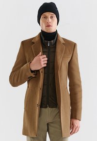 Brązowy wełniany płaszcz z klasycznym kołnierzem, guzikami i naszytymi kieszeniami, nałożony na ciemnozieloną puchową kamizelkę z zamkiem. Czarna, prążkowana czapka.