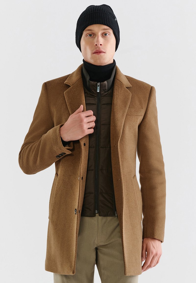 Brązowy wełniany płaszcz z klasycznym kołnierzem, guzikami i naszytymi kieszeniami, nałożony na ciemnozieloną puchową kamizelkę z zamkiem. Czarna, prążkowana czapka.