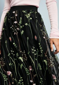 Jupe noire brodée avec une texture transparente ornée de motifs floraux verts, roses et blancs. La taille élastique améliore l'ajustement.