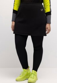 Zwarte sportieve skort met een gladde textuur, gecombineerd met zwarte leggings en neongroene sneakers. Heeft een logo op de tailleband.