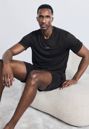 Αθλητικός άνδρας με μαύρο t-shirt και σορτς BOSS, καθισμένος σε μια υφασμάτινη, μπεζ μαξιλαροθήκη πάνω σε απλό φόντο.