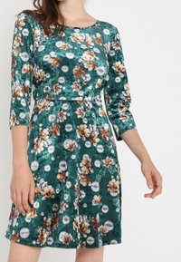 Blumenkleid aus grünem Samt mit drei Viertelärmeln, das weiße und orangefarbene Blumen sowie kreisförmige Muster aufweist.