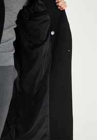 Cappotto nero con esterno liscio e fodera interna nera in raso. Presenta una tasca grande e una chiusura con bottoni.