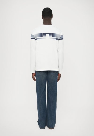 Λευκό πουκάμισο με μακριά μανίκια και οριζόντιο μπλε και γκρι αφηρημένο σχέδιο στο πάνω μέρος της πλάτης, συνδυασμένο με φαρδιά μπλε τζιν παντελόνια.