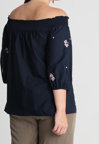 Femme portant un haut bleu marine à épaules dénudées avec broderies florales sur les manches et un pantalon beige, vue de dos et de côté.