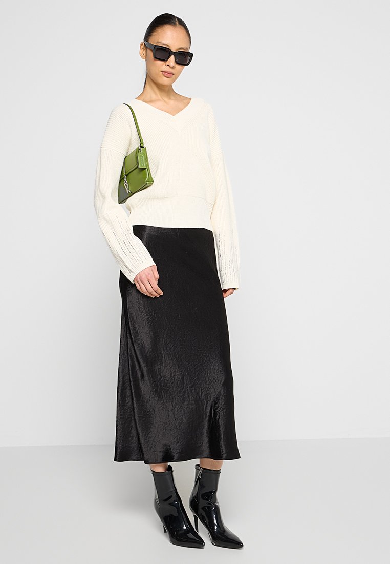 Maglione bianco a collo a V, gonna midi nera in satin, stivaletti neri alla caviglia e una piccola borsa verde. Gli accessori includono occhiali da sole neri.