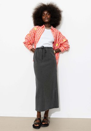 Femme aux cheveux afro portant une chemise rouge à carreaux par-dessus un top blanc, une longue jupe grise et des sandales noires, debout devant un fond blanc.