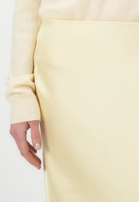 Light yellow, textured skirt with a side slit, paired with a soft beige sweater featuring ribbed cuffs. Hand displays subtle nail polish.