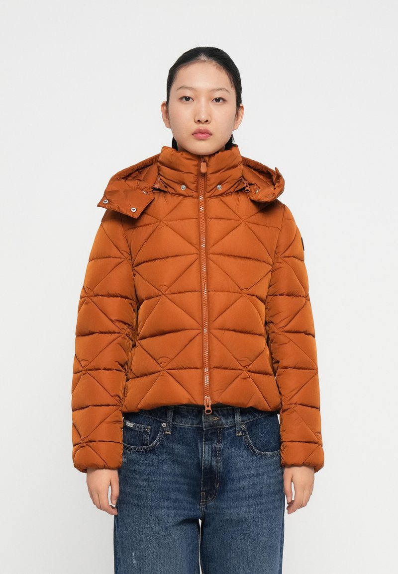 Pufferjakke i orange-brun, med høj krave, frontlynlås og diamantquiltet mønster. Kombineret med blå denimjeans.