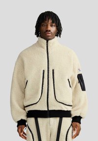 Giacca in pile crema con cerniera nera e dettagli Coordinati, colletto alto, due tasche frontali con cerniera, polsini a coste e pantaloni abbinati.