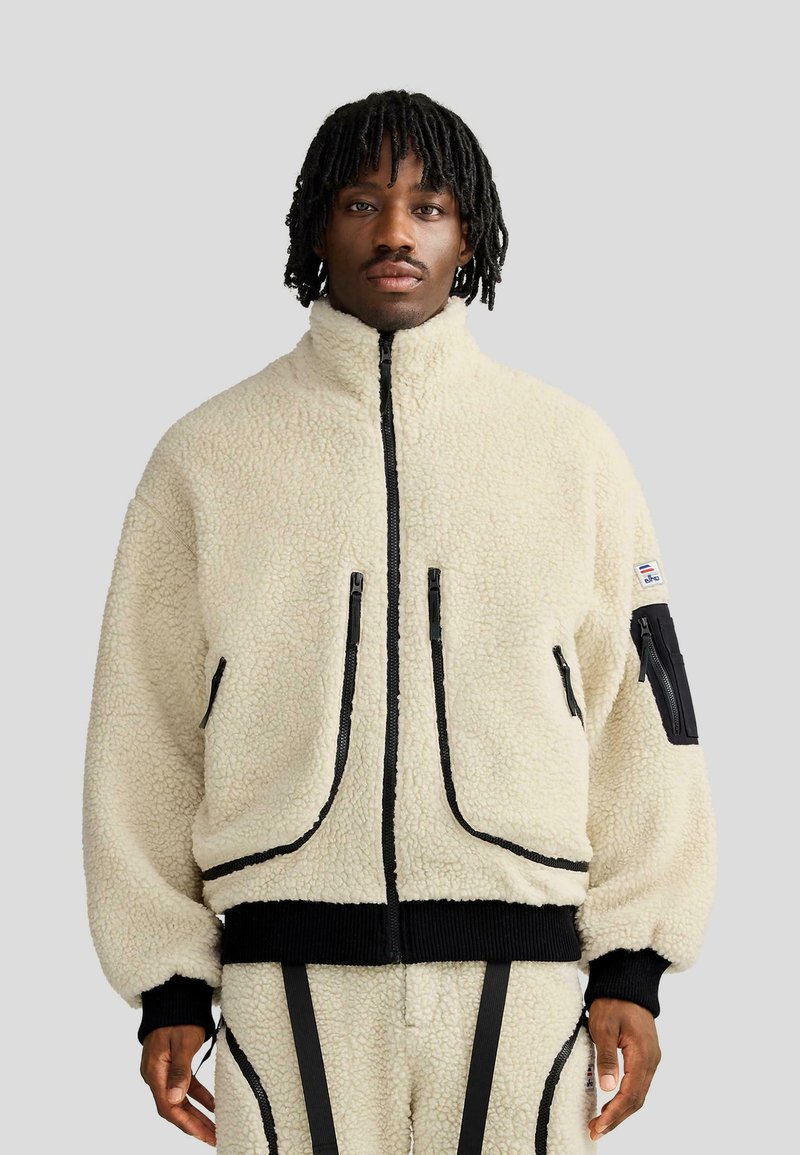 Giacca in pile crema con cerniera nera e dettagli Coordinati, colletto alto, due tasche frontali con cerniera, polsini a coste e pantaloni abbinati.