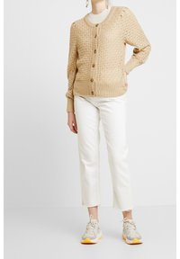 Cardigan beige en tricot avec motif diamant texturé, boutons en bois, poignets et ourlet côtelés, associé à un jean blanc court et des baskets grises.