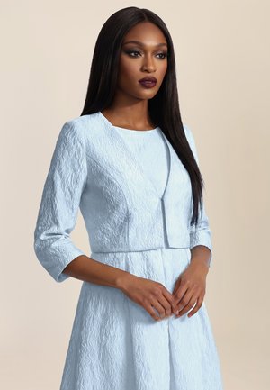 Woman with long straight hair wearing textured light blue jacket and matching skirt, standing with hands clasped, neutral background.