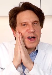 Uomo in camice da laboratorio con una camicia a righe blu, mani sul volto, che esprime coinvolgimento; lo sfondo chiaro mette in evidenza i tratti del viso.