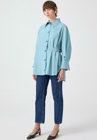 Camicia azzurro chiaro oversize con cintura in vita, abbinata a jeans a gamba dritta blu scuro e mule nere con tacco.