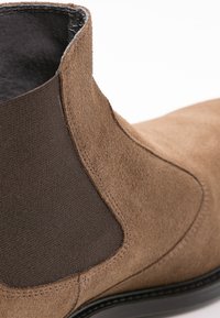 Botín de ante marrón con paneles laterales elásticos oscuros, punta redondeada y una suela negra lisa. Las superficies texturizadas contrastan con los detalles elegantes.