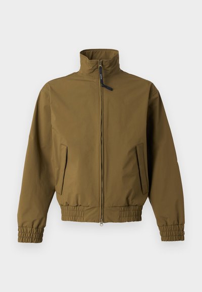 Barna cipzáras kabát magas gallérral, rugalmas mandzsettával és szegéllyel, két első zsebbel, egyszínű világos háttéren.