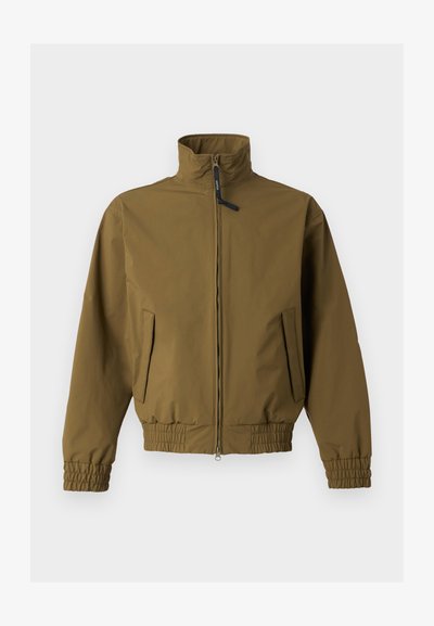 Barna cipzáras kabát magas gallérral, rugalmas mandzsettával és szegéllyel, két első zsebbel, egyszínű világos háttéren.