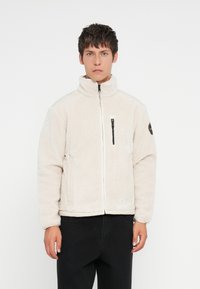 Giacca zip-up di colore crema sfocato con colletto alto, tasca sul petto con zip nera e tasche laterali. Tessuto morbido, vestibilità rilassata.