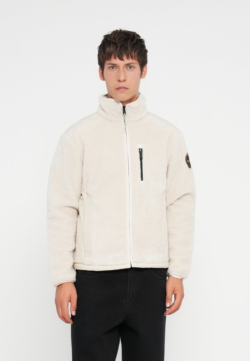 Giacca zip-up di colore crema sfocato con colletto alto, tasca sul petto con zip nera e tasche laterali. Tessuto morbido, vestibilità rilassata.