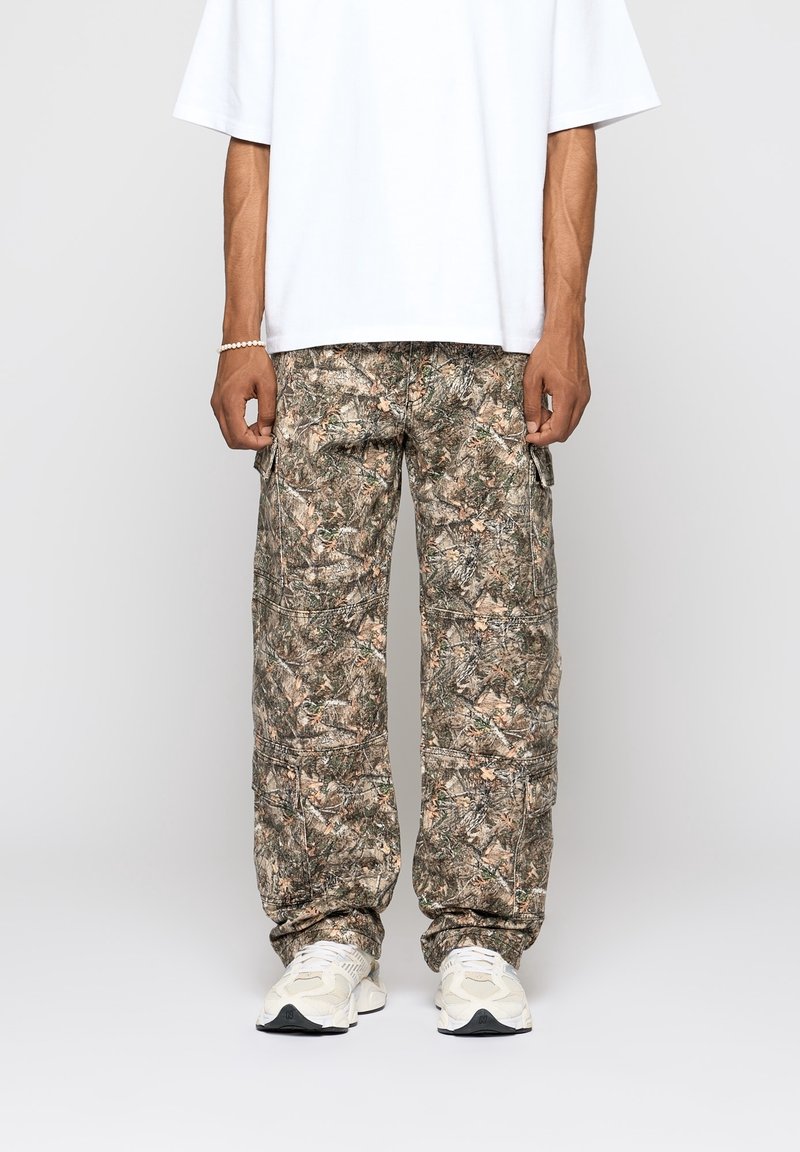 Homme portant un pantalon cargo camouflage, un t-shirt blanc oversize, des baskets blanches et un bracelet de perles au poignet gauche, debout devant un fond uni.