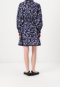 Femme portant une robe à manches longues bleue et blanche à motifs avec un ourlet festonné et des chaussures plates noires, vue de dos.