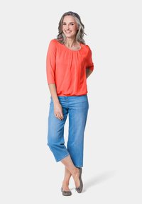 Korallefarbige Bluse mit Rundhalsausschnitt und Falten, kombiniert mit hellblauen, cropped Jeans und grauen Ballerinas. Glattes Material, lockere Passform.