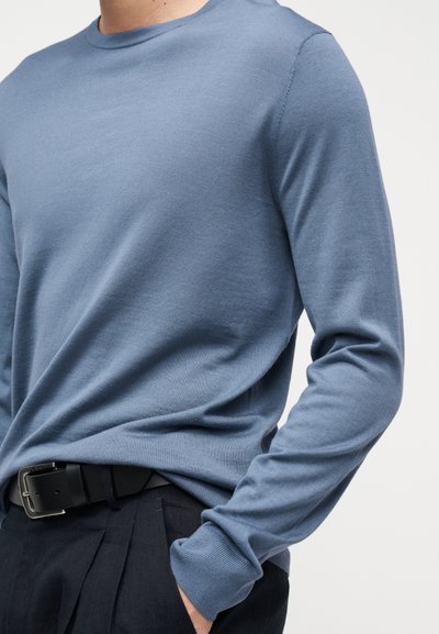 Άνδρας που φοράει εφαρμοστό, μακρυμάνικο μπλε πουλόβερ, βιδωμένο σε σκούρο παντελόνι με μαύρη δερμάτινη ζώνη, με το χέρι στην τσέπη μπροστά σε απλό φόντο.