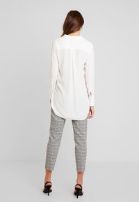 Blouse blanche à manches longues avec un ourlet arrondi et des poignets boutonnés, associée à un pantalon gris à carreaux longueur cheville et des sandales noires à talons.