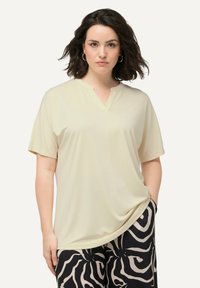 Blusa a maniche corte e scollo a V in tessuto beige chiaro. Texture morbida con vestibilità ampia; abbinata a pantaloni neri con un motivo bianco a vortice.