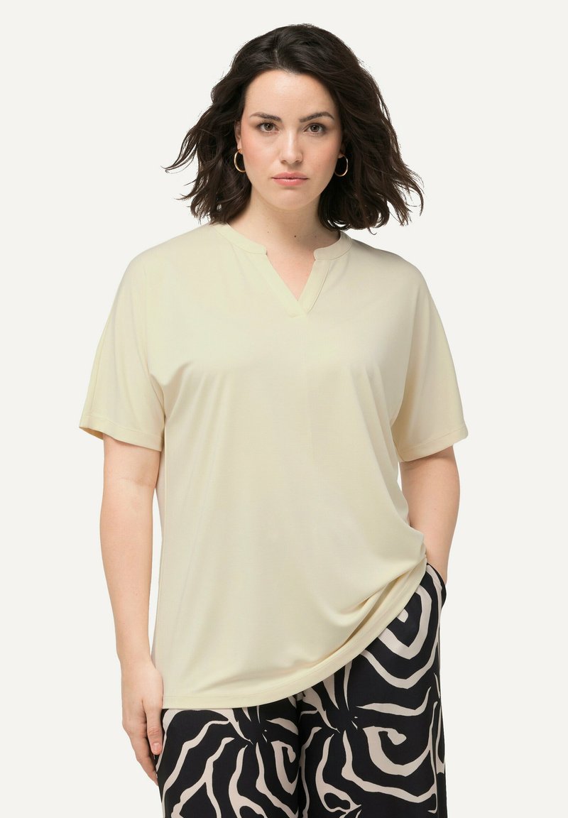 Blusa a maniche corte e scollo a V in tessuto beige chiaro. Texture morbida con vestibilità ampia; abbinata a pantaloni neri con un motivo bianco a vortice.