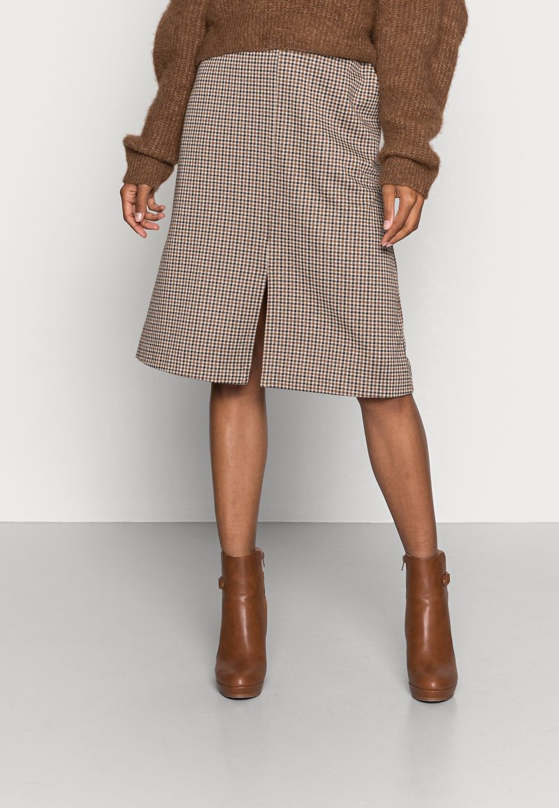 Personne portant un pull marron, une jupe à carreaux longueur genou avec fente devant et des bottines marron, debout devant un fond uni.