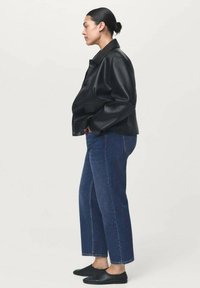 Veste en cuir noir avec un design à col, associée à un jean droit bleu et des chaussures noires à enfiler, vue de côté.