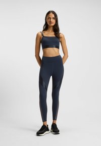 Woman standing with hands behind back wearing dark blue sports bra, matching high-waisted leggings, and black athletic shoes on plain background.