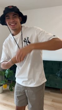 Bílá košile s krátkým rukávem a černým logem, šedé šortky a černý klobouk typu bucket s růžovým zdobením. Vnitřní prostředí se zelenou pohovkou a rostlinami.