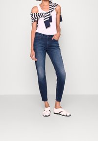 Persona que lleva jeans ajustados azul oscuro, una blusa blanca sin mangas, un suéter a rayas azul marino y blanco atado sobre los hombros, y sandalias blancas sin hebillas.