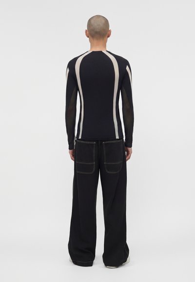 Άτομο με ξυρισμένο κεφάλι στέκεται με την πλάτη προς τα εμπρός φορώντας μαύρο μακρυμάνικο τοπ με λευκές κάθετες ρίγες και φαρδιά μαύρα παντελόνια με μεγάλες τσέπες.