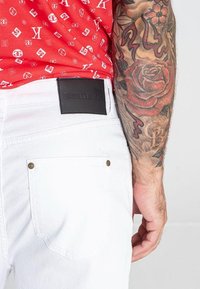 I jeans bianchi presentano cuciture minime, una tasca posteriore con un bottone dorato e una patch in pelle nera con la scritta "GIANNI KAVANAGH" in rilievo.