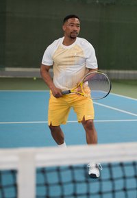 Tennis-Spieler, der ein Shirt mit weißem und gelbem Farbverlauf, gelbe Shorts und weiße Schuhe trägt, hält einen Schläger auf einem blauen Platz.