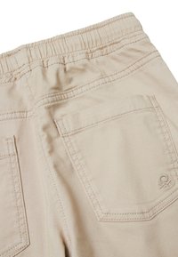 Béžové kalhoty s elastickým pasem, dvěma zadními kapsami a prošívanými detaily. Na spodní pravé kapse je znak.