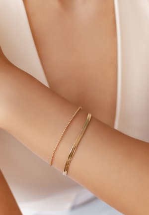Two gold bracelets on a wrist: one is a delicate beaded chain, and the other features a flat, textured design with a smooth finish.