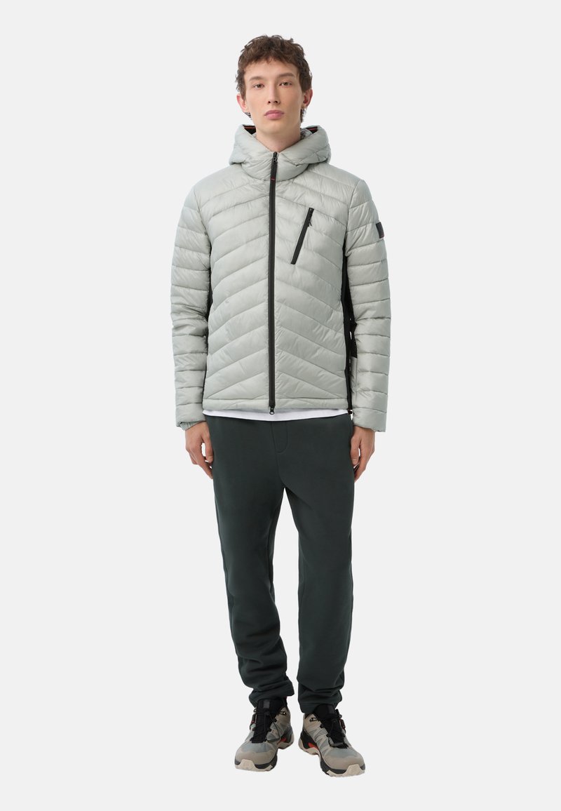 Helle graue, gesteppte Daunenjacke mit Kapuze, Frontreißverschluss und schwarzer Reißverschlusstasche, kombiniert mit dunkelgrünen Hosen und Sneakers.