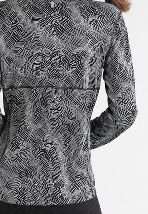 Vue de dos d'une personne portant une veste noire ornée de motifs abstraits ondulés blancs et à manches longues.