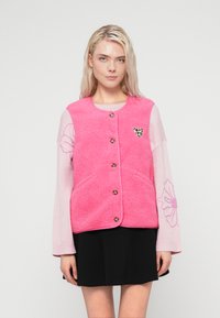 Ροζ γιλέκο sherpa με στρογγυλή λαιμόκοψη, πέντε καφέ κουμπιά και μια μικρή καρδιά με λεοπάρ μοτίβο. Φορεμένο πάνω από ένα ανοιχτό ροζ πουλόβερ με λουλουδένια σχέδια.