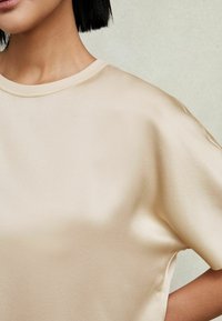Femme portant un haut court en satin beige lisse et brillant, visible des épaules jusqu'au milieu du torse sur un fond neutre.