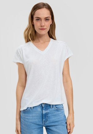 White v-neck t-shirt with short sleeves, featuring eyelet detail at shoulders. Soft, lightweight fabric with a relaxed fit. Paired with blue jeans.