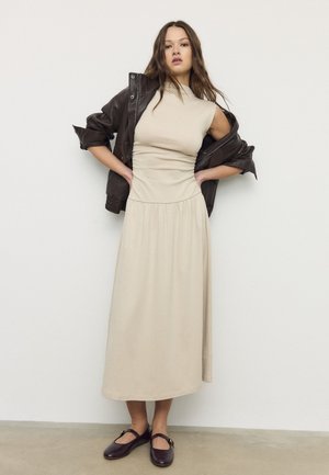 Žena u bež midi haljini bez rukava, s tamnoljubičastom kožnom jaknom spuštenom s ramena i tamnoljubičastim Mary Jane cipelama, stoji nasuprot bijelog zida.